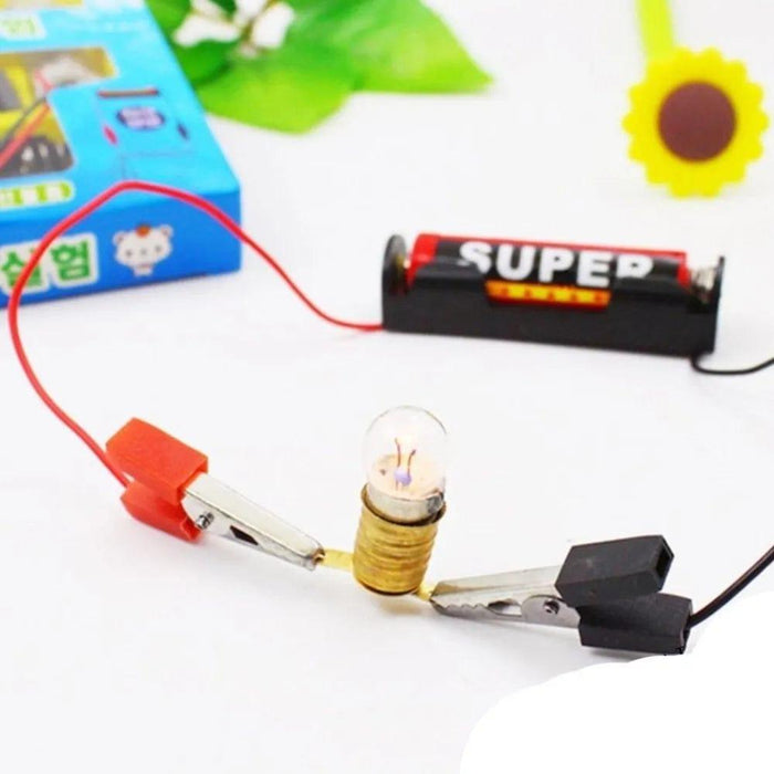 Small light bulb with wires and clips on a white background, with a battery and colorful packaging in the background.