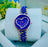 Blue heart-shaped watch with gold accents on a white cushion, with a blurred background