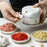 Person using a white handheld grinder with bowls of ground ingredients on a light surface.