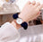 Hand wearing a watch with a navy blue scrunchie on a blurred indoor background