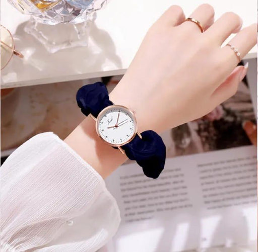 Hand wearing a watch with a navy blue scrunchie on a blurred indoor background